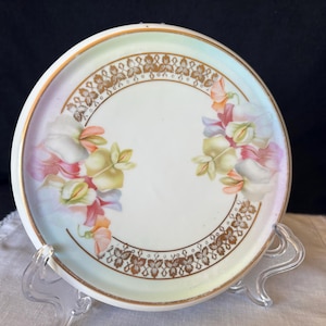 May include: A small, round, antique porcelain plate with a white center and a pastel-colored rim. The plate features a gold floral border and delicate floral designs in pink, yellow, and white. The plate is displayed on a clear stand.