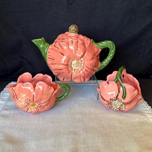 May include: A ceramic tea set featuring a teapot, creamer, and sugar bowl, all shaped like coral-colored poppy flowers. The teapot has a green handle and spout, and the sugar bowl has a matching green handle. The set is displayed on a white tablecloth.