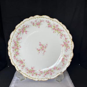 May include: A white porcelain plate with a gold rim and a delicate pink floral pattern. The plate has a scalloped edge and is perfect for serving a variety of dishes.