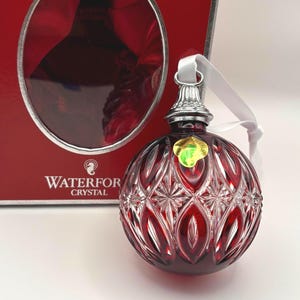 May include: A red Waterford Crystal ornament with a faceted diamond and leaf pattern, featuring a silver-toned metal cap and a white ribbon. The ornament is positioned in front of its red and silver box with the Waterford Crystal logo printed on it.