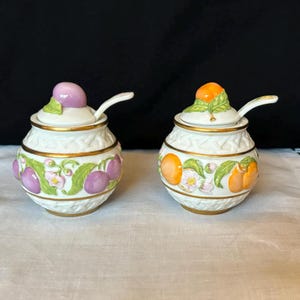 May include: Two white ceramic condiment jars with lids and spoons. The jars are decorated with a woven pattern and fruit designs. One jar has purple plums and the other has orange peaches.