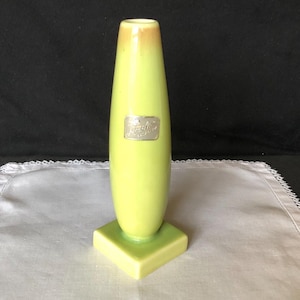 May include: A lime green ceramic vase with a square base and a slender, elongated body. The top of the vase has a light brown hue. A silver rectangular label is affixed to the front of the vase.