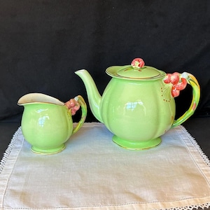 May include: A vintage green ceramic tea set, comprising a teapot and a creamer. The teapot has a lid with a floral knob and a curved spout. Both pieces feature gold trim and handles decorated with red and pink floral accents. The set is presented on a white cloth.