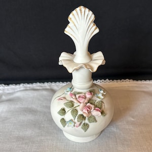 May include: A white ceramic perfume bottle with a decorative floral design. The bottle features a round base, a bulbous body, and a tall, ornate stopper with gold accents. Pink roses and green leaves adorn the body, with small blue flowers.