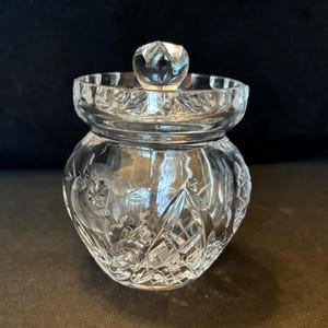 May include: A clear, cut-glass jar with a lid. The jar has a round body with etched floral and star designs. The lid has a round knob. The jar is likely used for storing small items or as a decorative piece.