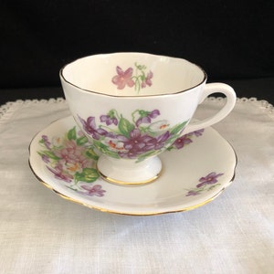 May include: A white teacup and saucer with a floral pattern of purple and pink flowers. The cup and saucer have a gold trim.