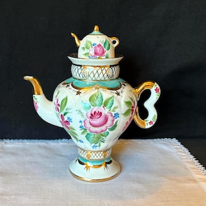 May include: A white porcelain teapot featuring a smaller teapot-shaped lid, decorated with hand-painted pink roses, green leaves, and blue accents. The piece includes gold-painted trim on the spout, handle, base, and a lattice pattern band.