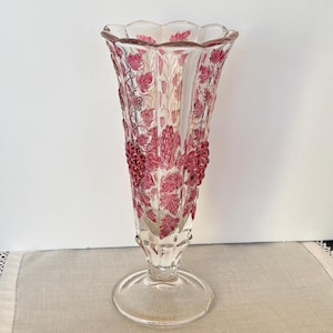 May include: A clear glass vase with a flared, scalloped rim. The vase is decorated with a red grape and leaf design. The vase has a round base and a long, slender stem. The vase is approximately 10 inches tall.