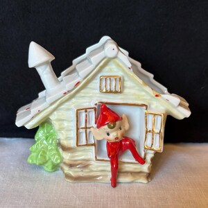 May include: A ceramic Christmas elf figurine peeking out of a small house. The house is painted in cream and white with gold trim, a chimney, and a green tree. The elf wears a red outfit and hat.