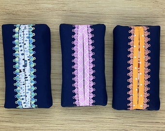 Könnte beinhalten: Drei dunkelblaue Taschentuchbox-Bezüge mit verschiedenen gemusterten Besätzen. Der Besatz des linken Bezugs ist ein weiß-blaues geometrisches Muster, der mittlere Bezug hat ein rosa-weißes Blumenmuster und der rechte Bezug hat ein orange-weißes Punkte-Muster.