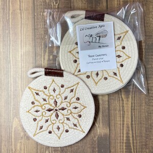 Può includere: Due sottobicchieri rotondi in corda con un motivo a stella floreale. I sottobicchieri sono bianco sporco con ricami marroni e dorati. Ogni sottobicchiere ha un anello e un inserto in pelle marrone. Il testo "Lil Creative Apis" e "Rope Coasters Floral star" è incluso.