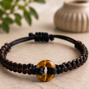 Puede incluir: Una pulsera con una cuenta central circular de ojo de tigre, flanqueada por un patrón trenzado de cordón negro y marrón. La pulsera incluye un cierre deslizante ajustable hecho del mismo cordón negro para un ajuste seguro.