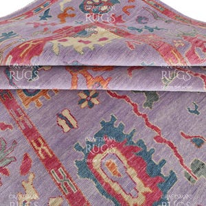 Op de afbeelding: Een opgevouwen paars tapijt met een gedetailleerd patroon in tinten rood, teal, oranje en crème. Het tapijt heeft een herhalend ontwerp met bloemen en geometrische elementen. De woorden "CRAFTSMAN RUGS DECORATE HOME" staan op het tapijt.