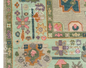 Può includere: Primo piano di una sezione di un tappeto decorativo con un motivo floreale. Il tappeto presenta uno sfondo verde chiaro con un bordo di disegni floreali verdi, rosa e viola. Il testo "CRAFTSMAN RUGS DECOR THE HOME" è visibile.