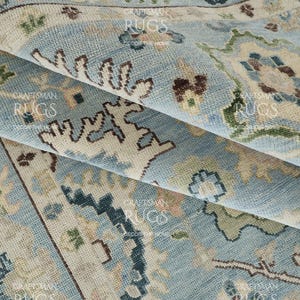 Pu&ograve; includere: Primo piano di un tappeto blu chiaro piegato con un motivo tradizionale intricato. Il tappeto presenta accenti beige, verdi e marroni, creando una superficie dettagliata e strutturata. Il design del tappeto include elementi floreali e geometrici.