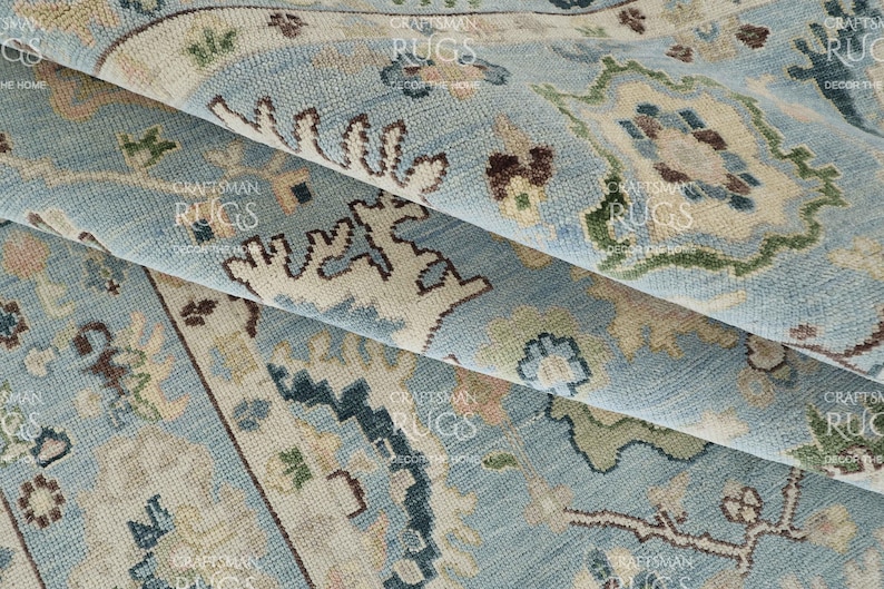 Pu&ograve; includere: Primo piano di un tappeto blu chiaro piegato con un motivo tradizionale intricato. Il tappeto presenta accenti beige, verdi e marroni, creando una superficie dettagliata e strutturata. Il design del tappeto include elementi floreali e geometrici.
