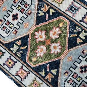 Op de afbeelding: Close-up van een kleurrijk tapijt met een ingewikkeld geometrisch patroon. Het tapijt heeft een centraal groen gedeelte met witte bloemmotieven, omgeven door marineblauwe, lichtblauwe en crèmekleurige randen. Het tapijt is een decoratief woondecoratie-item.
