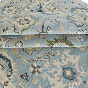 May include: Close-up of a folded, light blue rug with an intricate floral pattern in beige, green, and brown. The rug has a distressed, vintage look, with visible texture and detail. The rug is labeled "Craftsman Rugs, Decor the Home."
