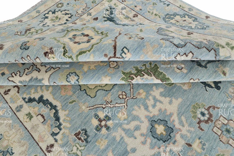 Pu&ograve; includere: Primo piano di un tappeto blu chiaro piegato con un intricato motivo floreale in beige, verde e marrone. Il tappeto ha un aspetto invecchiato e vintage, con texture e dettagli visibili. Il tappeto &egrave; etichettato "Craftsman Rugs, Decor the Home."