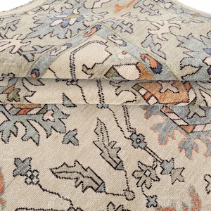 May include: A folded area rug featuring a light beige background with a repeating floral and geometric pattern in shades of muted blue, burnt orange, and sage green. The textile displays intricate woven details and a soft, textured surface.