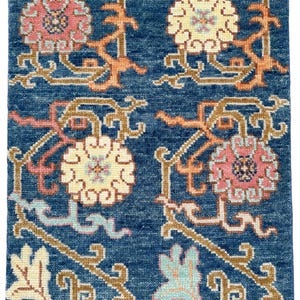 以下が含まれることがあります： 青いラグのクローズアップ。繰り返し模様の花柄。ピンク、黄色、クリーム色の様式化された花のデザインで、オレンジと水色のアクセントがあります。ラグはテクスチャード加工されています。