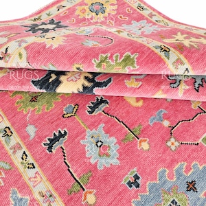 Op de afbeelding: Een opgevouwen roze tapijt met een gedetailleerd bloemenpatroon. Het tapijt heeft een combinatie van kleuren, waaronder roze, blauw, groen en geel. Het ontwerp bevat bloemen en geometrische elementen.
