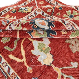 May include: Close-up of a folded red rug with intricate floral and geometric patterns. The rug features a mix of colors including red, blue, green, and cream. The design is detailed and appears to be of high quality.