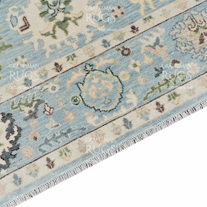 Pu&ograve; includere: Primo piano di una sezione di un tappeto azzurro con un intricato motivo floreale. Il tappeto presenta un bordo color crema con disegni dettagliati in verde, marrone e blu. Il bordo del tappeto ha una frangia.