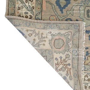 May include: Close-up of a section of a beige and blue patterned rug. The rug features floral and geometric designs in shades of blue, beige, and pink. The rug has a fringed edge and a folded corner, revealing the intricate details.