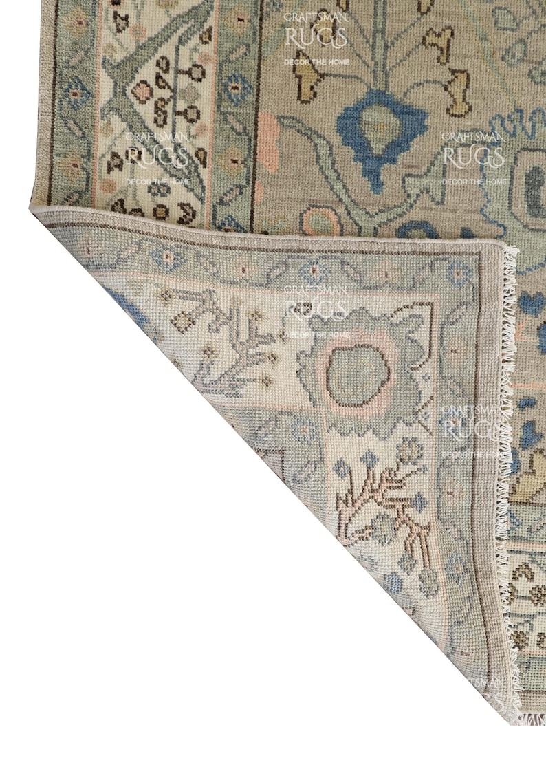 May include: Close-up of a section of a beige and blue patterned rug. The rug features floral and geometric designs in shades of blue, beige, and pink. The rug has a fringed edge and a folded corner, revealing the intricate details.