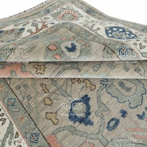 以下が含まれることがあります： ベージュ、ブルー、ピンクの色合いで詳細な花柄が施された、折りたたまれたラグのクローズアップ。 ラグは使い古されたヴィンテージの外観です。「CRAFTSMAN RUGS DECOR THE HOME」というテキストが表示されています。