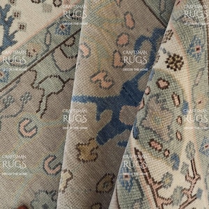 May include: Close-up of a folded area rug with a detailed, vintage design. The rug features a muted colour palette of beige, blue, and pink, with intricate floral and geometric patterns. The rug's texture is visible, suggesting a woven construction.