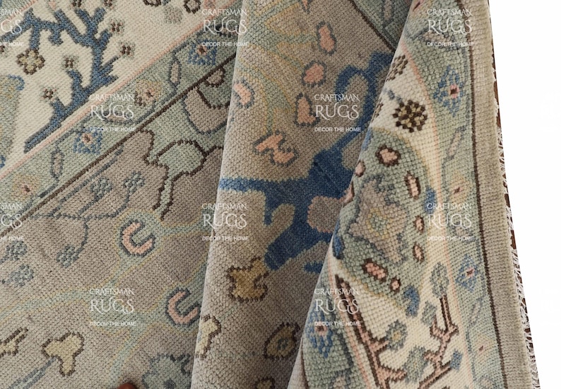 May include: Close-up of a folded area rug with a detailed, vintage design. The rug features a muted colour palette of beige, blue, and pink, with intricate floral and geometric patterns. The rug's texture is visible, suggesting a woven construction.