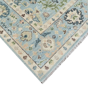 Pu&ograve; includere: Un angolo di un tappeto azzurro con un motivo floreale dettagliato. Il tappeto presenta un bordo color crema con piccoli accenti floreali nei toni del rosa, verde e marrone. Il design include un medaglione centrale ed elementi angolari.