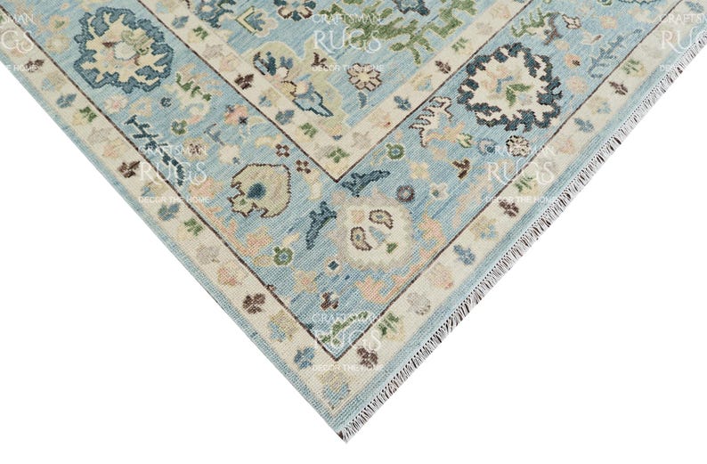 Pu&ograve; includere: Un angolo di un tappeto azzurro con un motivo floreale dettagliato. Il tappeto presenta un bordo color crema con piccoli accenti floreali nei toni del rosa, verde e marrone. Il design include un medaglione centrale ed elementi angolari.