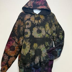 May include: A black hooded sweatshirt featuring a repeating sunflower pattern in various muted tones including pink, orange, yellow, green, and blue. The long-sleeve pullover includes a front kangaroo pocket, a drawstring hood, and ribbed cuffs.
