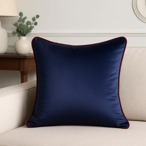 Pode incluir: Uma almofada decorativa quadrada azul marinho com acabamento bordô está sobre um sofá creme. A almofada tem uma textura lisa, semelhante à de cetim. O fundo inclui uma mesa lateral de madeira, uma lâmpada e um vaso com vegetação.