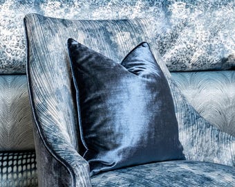 May include: A blue velvet pillow sits on a blue patterned armchair. The armchair has a high back and a curved shape.