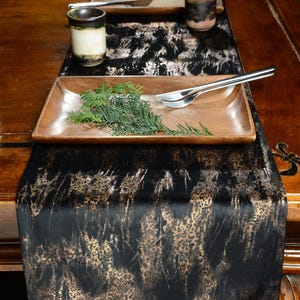 May include: Black velvet table runner with a gold metallic print of abstract shapes. The runner is displayed on a wooden table with a wooden tray, two ceramic mugs, and silver cutlery.