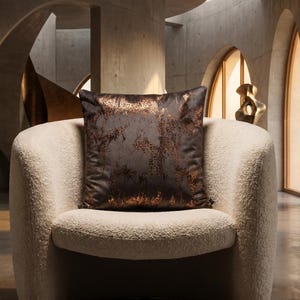May include: A decorative square pillow with a dark brown and copper-colored spotted pattern sits on a cream-colored, rounded armchair. The pillow's design resembles a leopard print. The setting is a modern interior with concrete walls and arched windows.