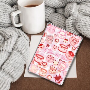 May include: A clear tablet case featuring a pink and white checkered pattern with various cake illustrations. The case has the text "Let them eat cake" in a cursive font. A white mug with coffee sits next to the case, with a gray knitted blanket in the background.