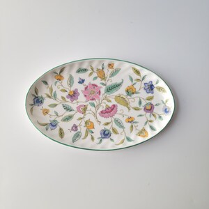 May include: Oval serving platter with a white background and a floral design. The flowers are pink, purple, blue, and yellow, with green leaves and a green border. The platter is a decorative piece.