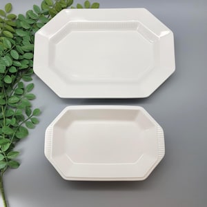 May include: Two white, octagonal serving platters. The larger platter is at the top, and the smaller one is below. Both have a raised, textured border. The platters are on a gray surface, with green leafy sprigs in the upper left corner.