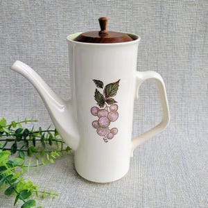 May include: White ceramic coffee pot with a wooden lid and handle. The pot features a grape cluster design in pink and green. The spout is long and curved. A vintage kitchenware item.