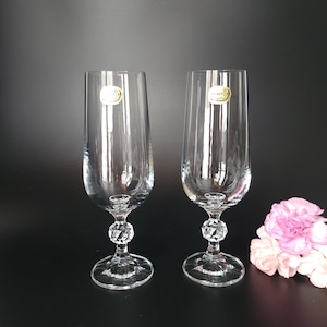 May include: Two clear crystal champagne flutes with a faceted crystal ball stem and round base. Each glass has a gold sticker. A pink flower is in the background. The glasses are on a black surface.