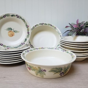 May include: A collection of off-white ceramic dishes with fruit and leaf designs. The set includes plates, bowls, and a serving dish. The plates have a fruit and berry border, and some feature a central orange illustration. A decorative floral arrangement is in the background.