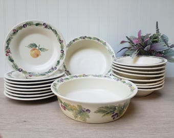 May include: A collection of off-white ceramic dishes with fruit and leaf designs. The set includes plates, bowls, and a serving dish. The plates have a fruit and berry border, and some feature a central orange illustration. A decorative floral arrangement is in the background.