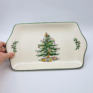 May include: A rectangular cream-colored ceramic serving tray with a green rim. The center features a decorated Christmas tree with colorful ornaments and gifts underneath. Sprigs of holly are painted on the left and right sides.