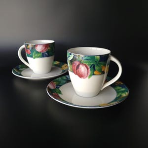 May include: Two white ceramic teacups and saucers. The cups and saucers have a colorful fruit and leaf design. The cups have a handle and are sitting on matching saucers. The background is black.