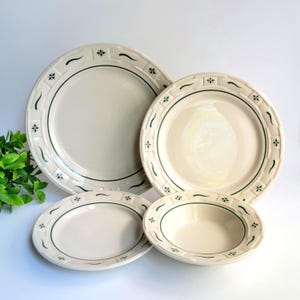 May include: A set of four off-white ceramic dishes featuring a dark green rim pattern with alternating geometric squares and curved lines. The collection includes two large dinner plates and two smaller bowls, all sharing a consistent design.